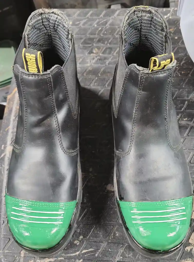 A pair of worn black work boots with bright green toe caps and yellow pull-tabs sit on a textured black surface. The boots…