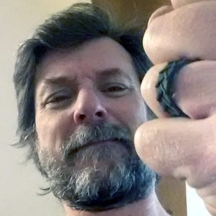 A close-up selfie of a middle-aged man with a graying beard and dark hair, looking directly at the camera with a slightly…