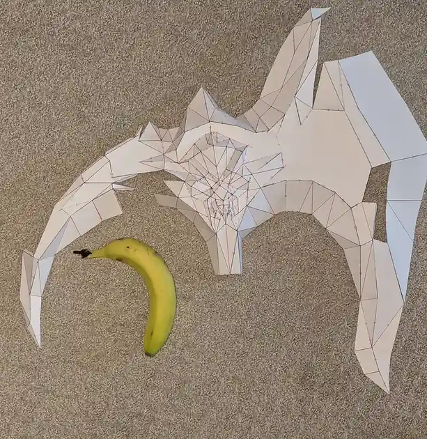 A flat, white paper cutout of a stylized bat with geometric facets and red fold lines lies on a beige carpet, next to a ripe…