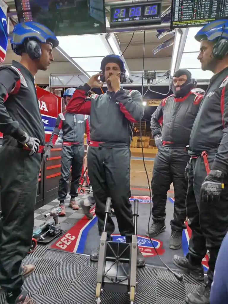 A group of race team members in racing suits and helmets stand in a pit garage, looking at a digital timer showing 21:35:15…