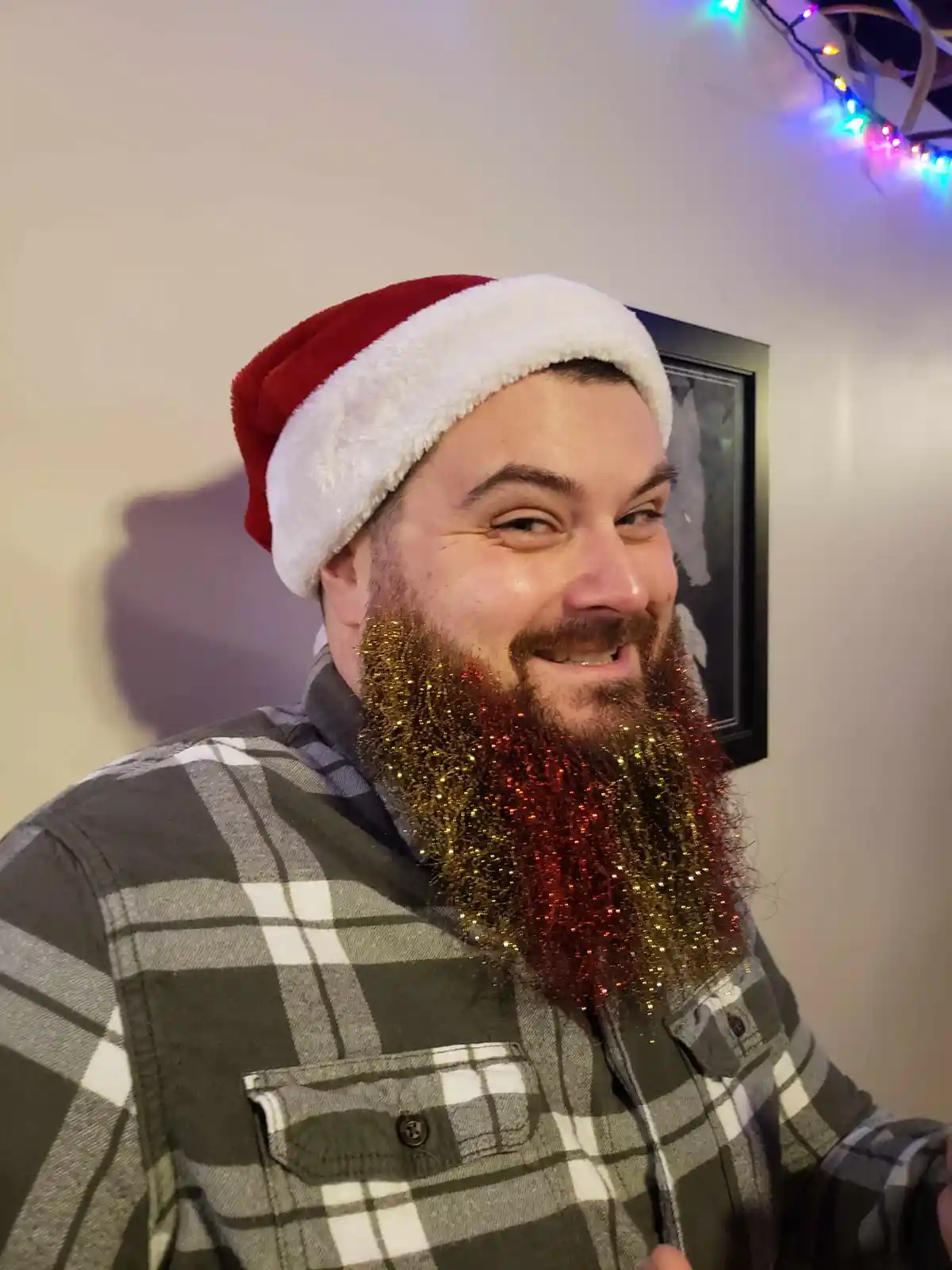A man wearing a red and white Santa hat and a plaid shirt smiles at the camera while his beard is covered in red and gold…