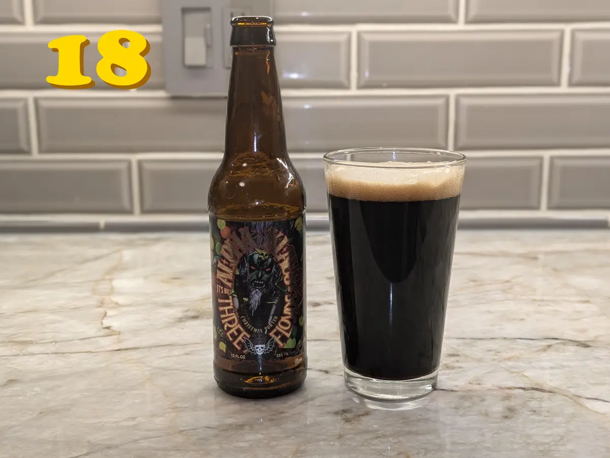 A brown beer bottle labeled 'It's Not All Three Christmas Porter' sits next to a full glass of dark beer on a marble…