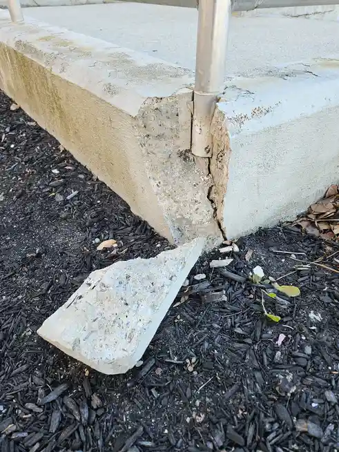 A close-up photo shows a large crack in a concrete structure, with a broken piece of concrete lying on dark mulch below. A…
