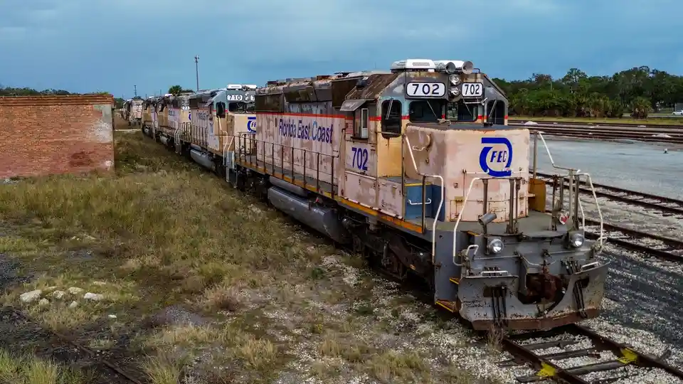 A row of rusty, abandoned Florida East Coast Railway locomotives sits idle on tracks next to overgrown grass and a brick…