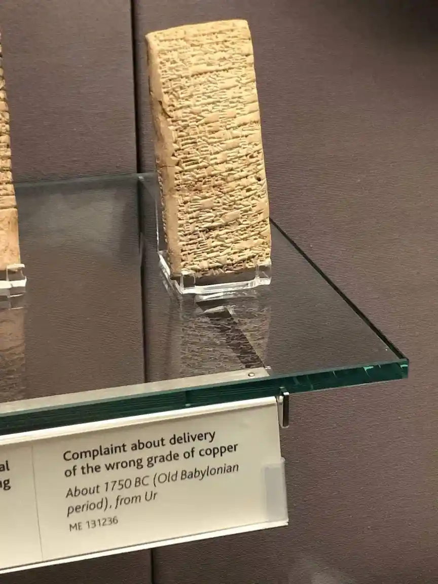 A close-up photograph of an ancient cuneiform tablet displayed in a museum case, with a label describing it as a 'Complaint…