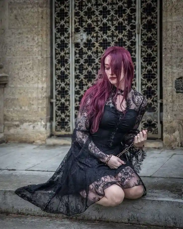 A woman with long magenta hair, wearing a black lace dress and choker, kneels on stone steps in front of an ornate metal…