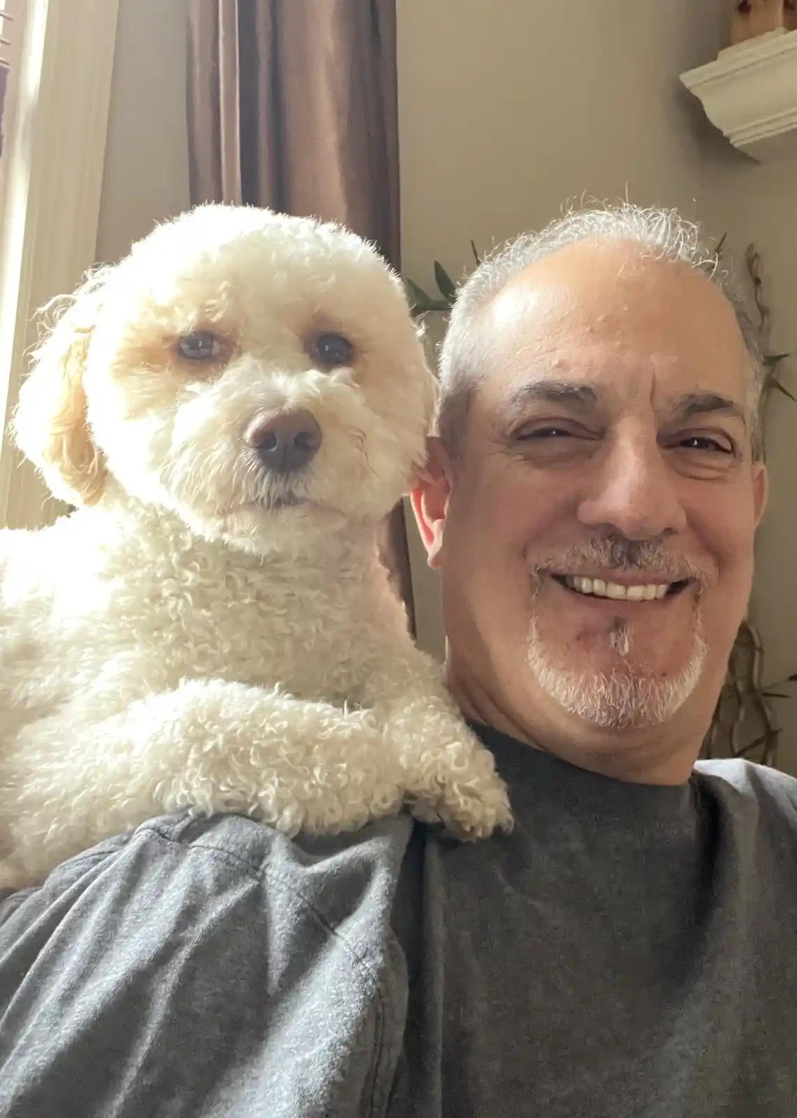 A man with gray hair and a goatee smiles broadly while holding a fluffy white dog on his shoulder. The dog, with curly fur…