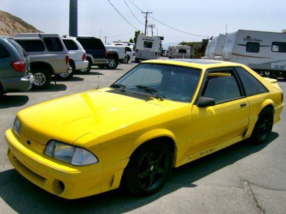 a bright yellow 1990s ford mustang coupe is parked in a lot with other vehicles, including suvs and rv's, under a clear sky…