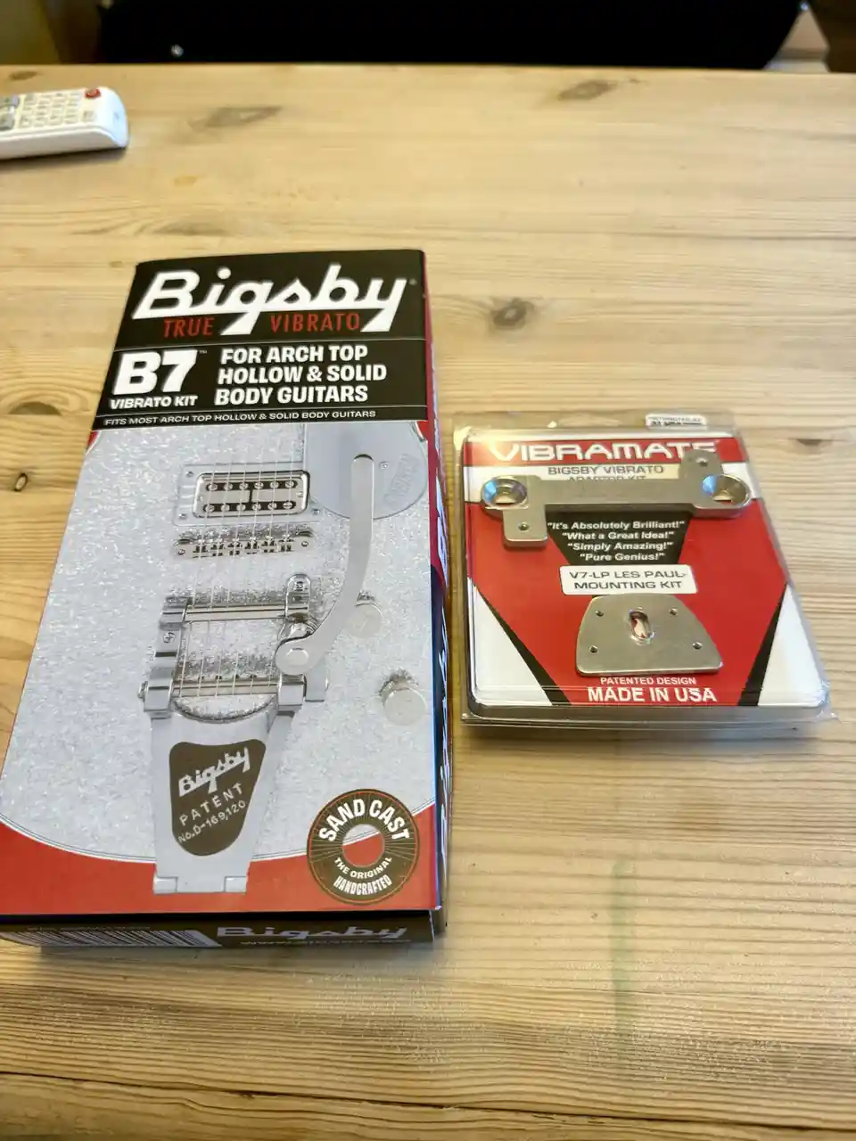 Two guitar vibrato kits sit side-by-side on a wooden table: a Bigsby True Vibrato B7 kit in its box and a Vibramate Bigsby…