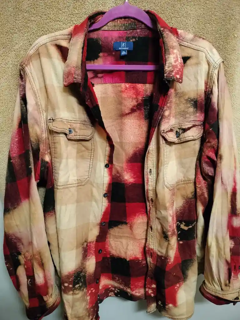 A red, black, and beige plaid flannel shirt hangs on a purple hanger against a beige carpeted wall. The shirt is visibly…