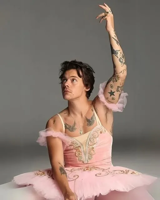 A man with visible tattoos on his arms and chest is seated, wearing a pink ballet tutu and bodice with ruffled sleeves. He…