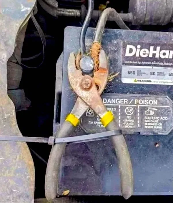 A pair of rusty, yellow-handled pliers is strapped to a car battery with a black zip tie, with the battery's warning label…