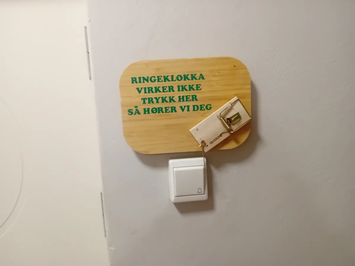 A white doorbell button is mounted on a wall, with a wooden sign above it in green text that reads 'RINGEKLOKKA VIRKER IKKE…