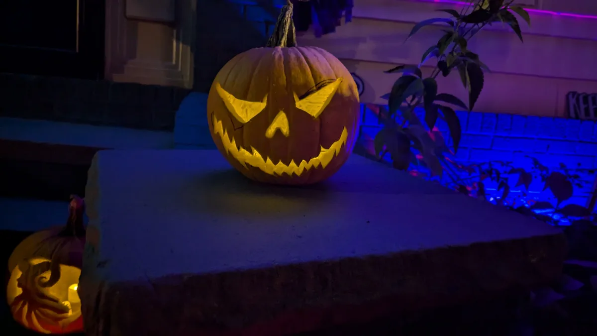 A carved jack-o'-lantern with a menacing, angular face sits on a stone step at night, illuminated from within by a warm…