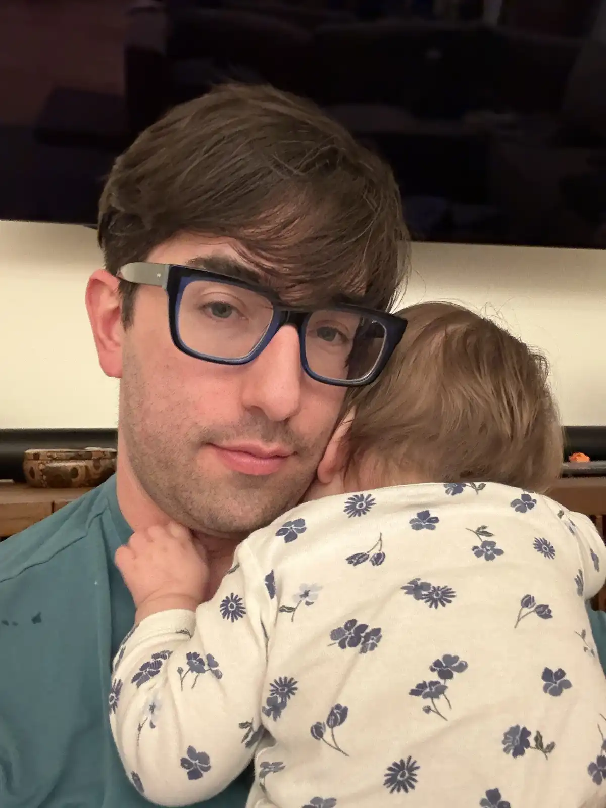 A man with dark hair and thick-rimmed glasses looks directly at the camera with a slightly weary or resigned expression…
