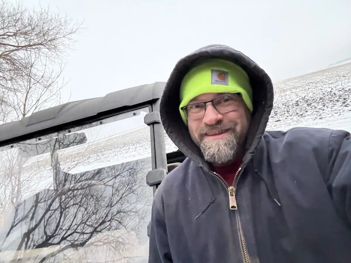 A man with glasses and a beard, wearing a bright green Carhartt beanie under a dark hooded jacket, smiles at the camera…