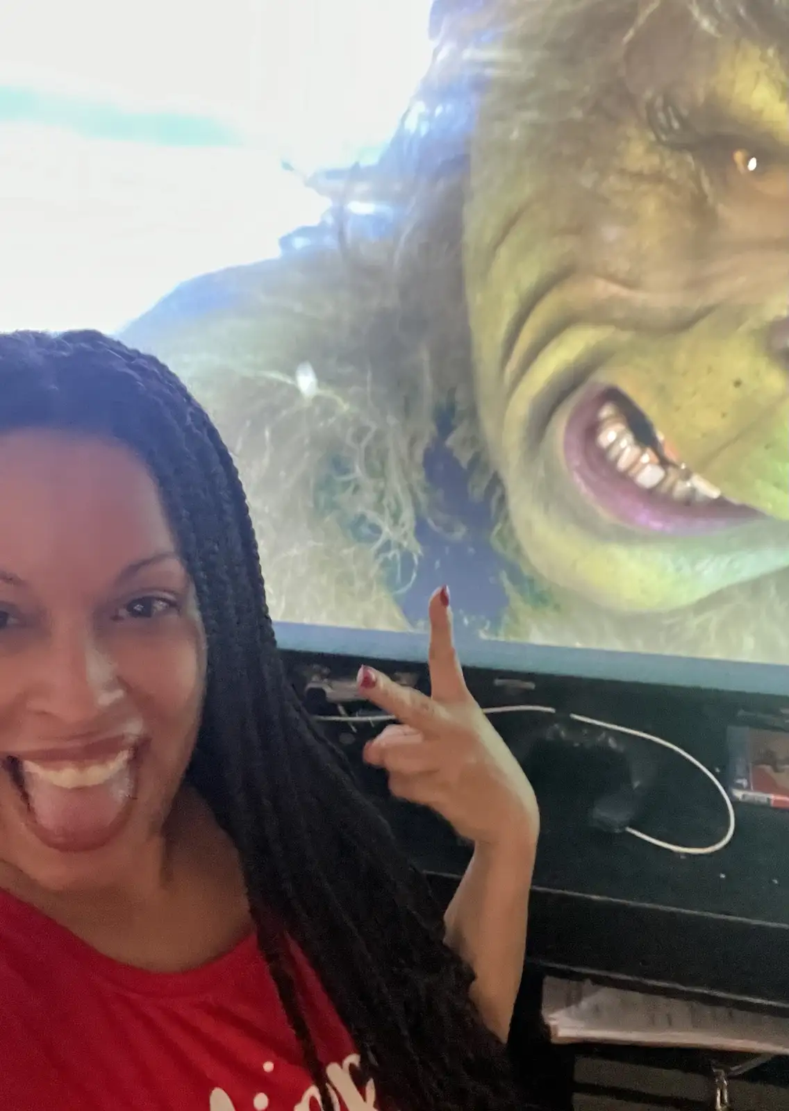 A woman with long dark braids and a red shirt poses for a selfie, sticking her tongue out and making a peace sign with her…