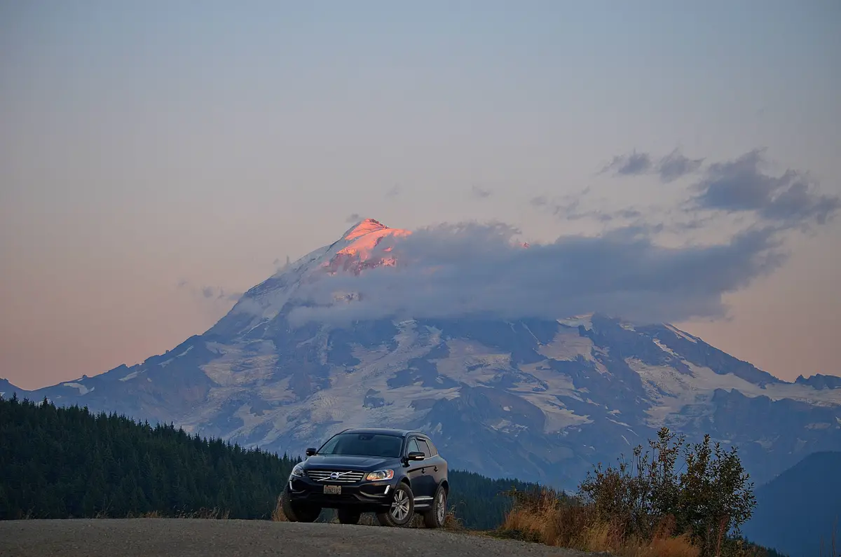 A dark blue Volvo SUV is parked on a gravel road in front of a majestic snow-capped mountain partially obscured by clouds…