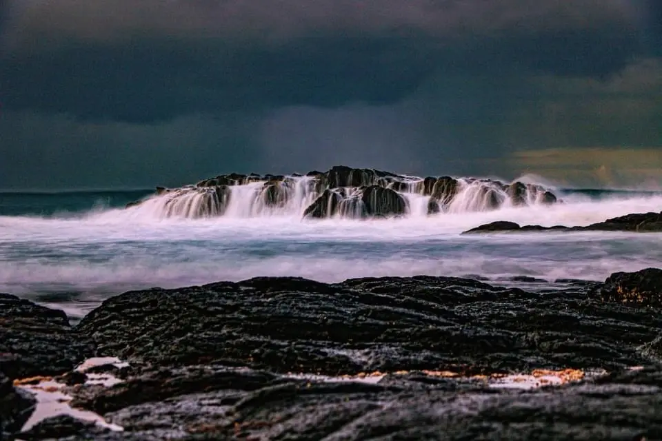 A long-exposure photograph of crashing ocean waves over dark volcanic rocks under a stormy sky, with the overlaid text 'i'm…