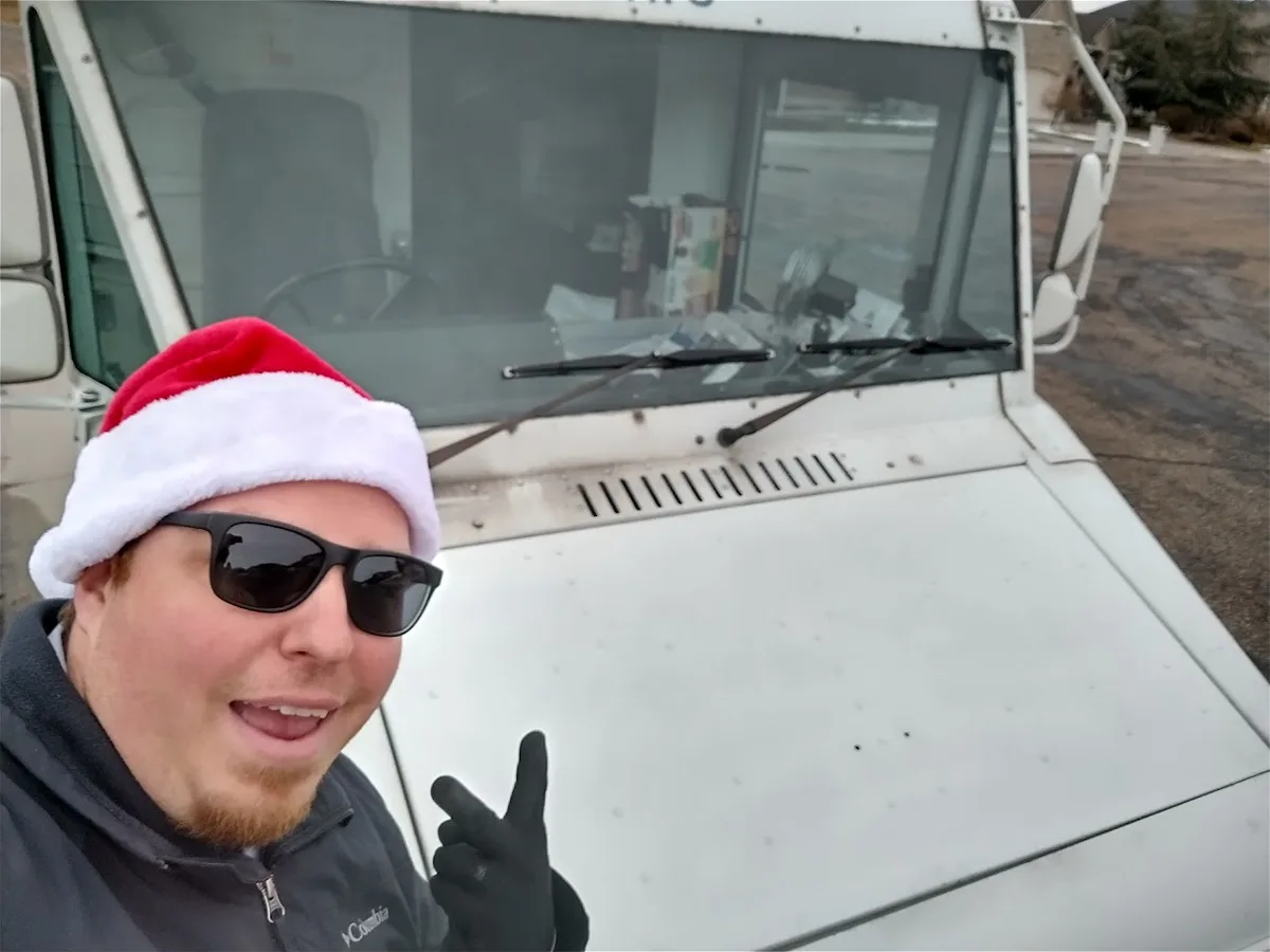 A man wearing a Santa hat, sunglasses, and black gloves stands in front of a white snow plow truck, pointing at the camera…