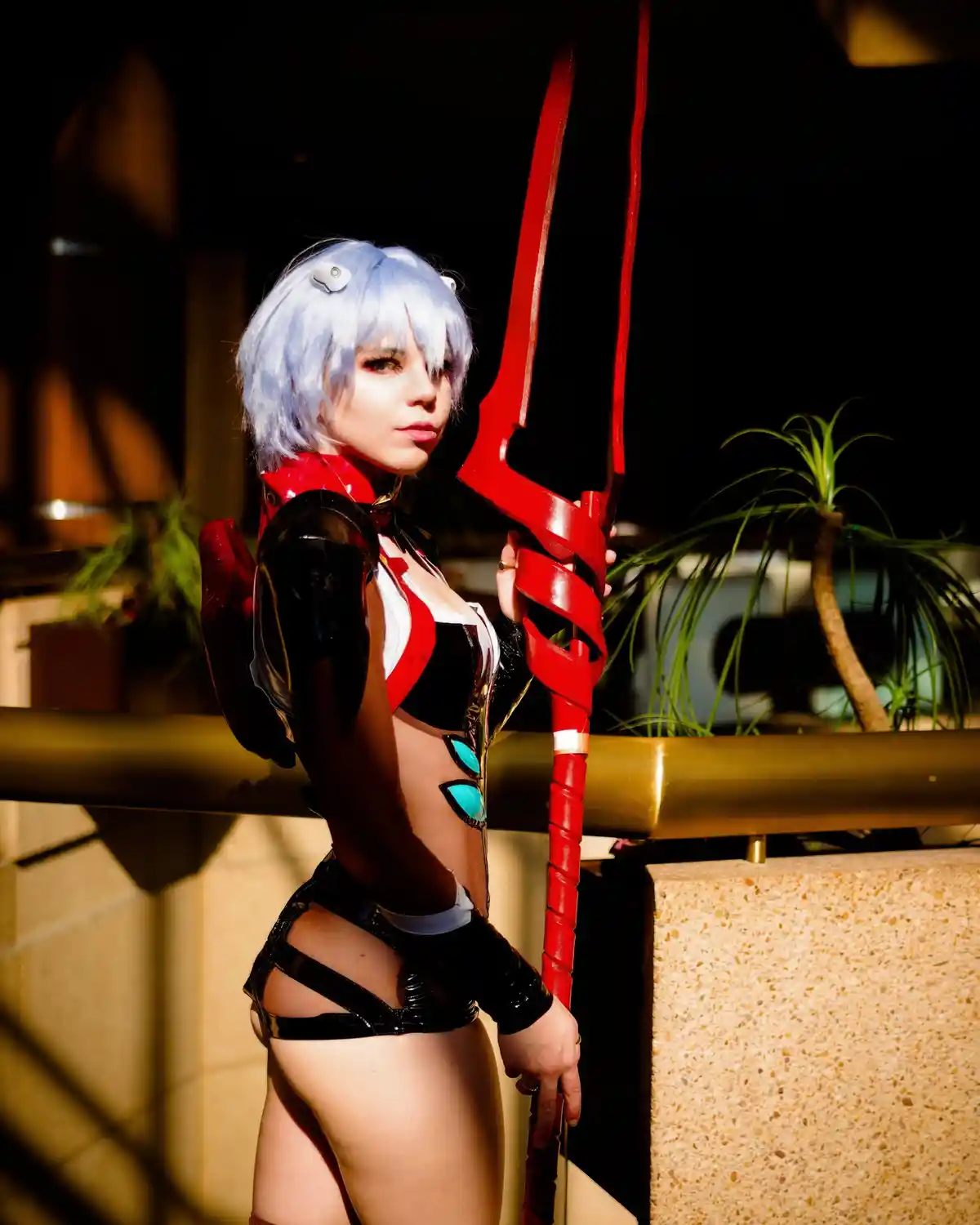 A person in a cosplay outfit resembling a character with short blue hair and red and black armor, holding a large red…