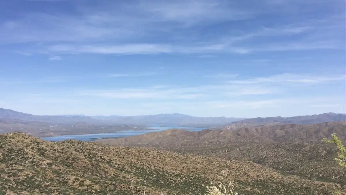 A wide-angle photo shows a vast desert landscape with rolling brown hills in the foreground and a large blue lake nestled…