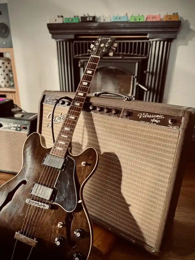 A vintage Gibson electric guitar with a worn, dark finish leans against a Fender Vibrosonic amplifier in a warmly lit room…