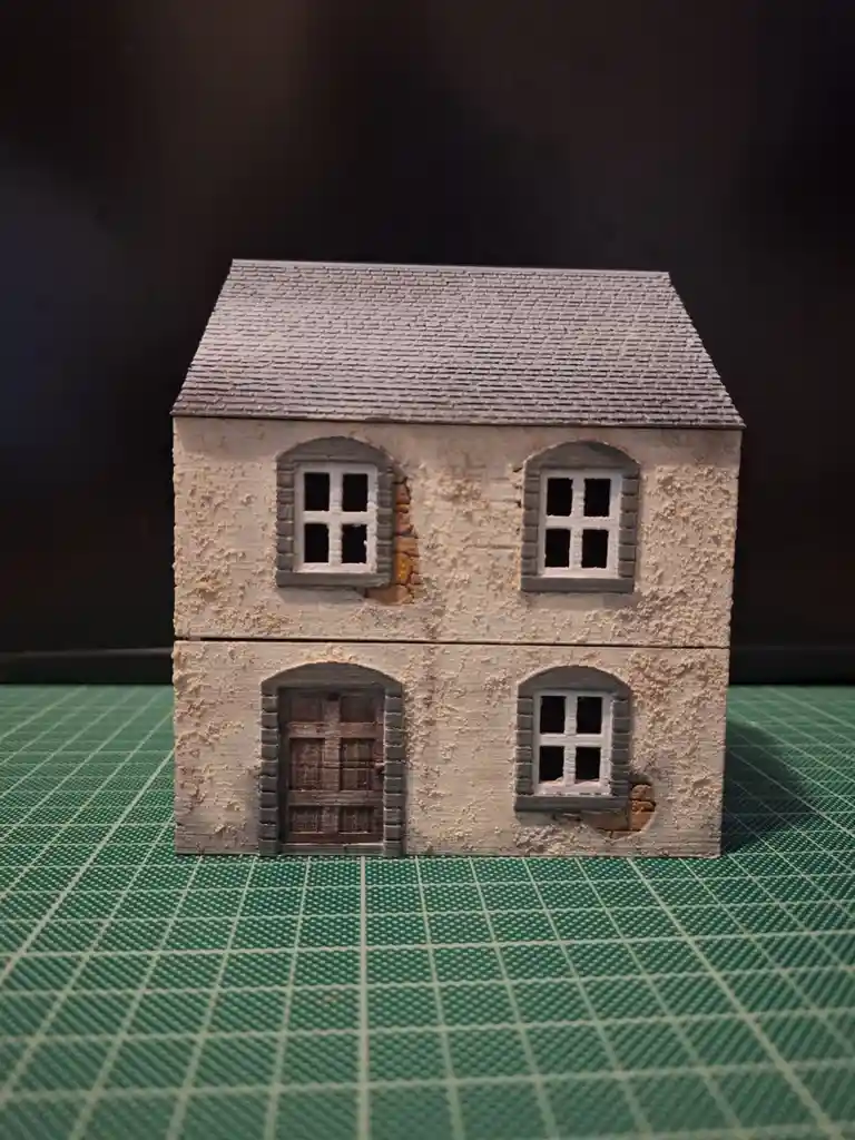 A small, two-story miniature house model with a gray roof, textured beige walls, and arched windows and door sits on a green…