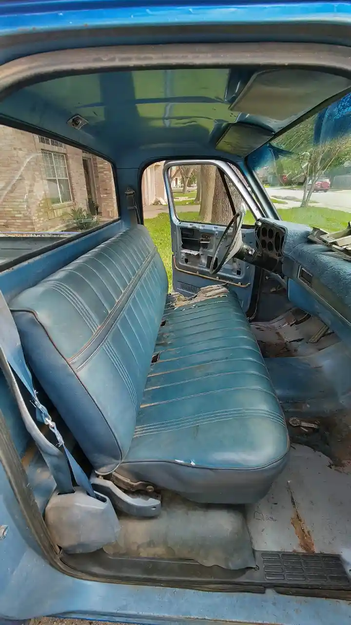 The image shows the interior of a blue vintage truck with a worn, torn blue vinyl bench seat. The driver's side door is…