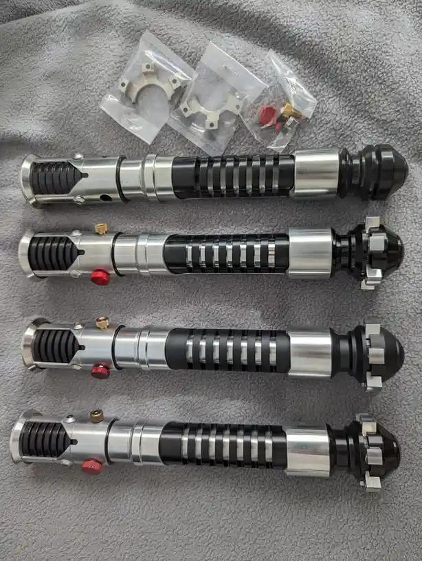 Four disassembled lightsaber hilt components lie horizontally on a gray towel, each with a metallic finish and black grip…