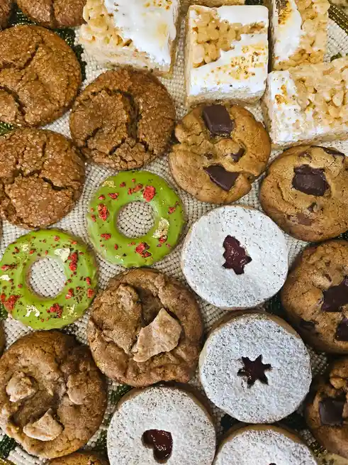 An overhead shot displays a colorful assortment of holiday-themed baked goods, including chocolate chip cookies, rice…