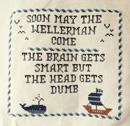 A close-up of a cross-stitched textile with a decorative border, featuring two small stitched boats at the bottom and text…