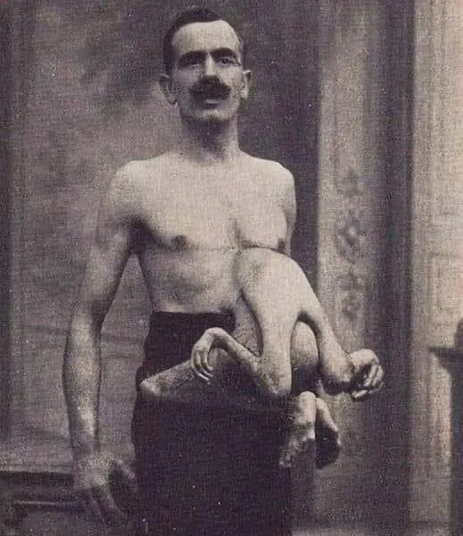 A black-and-white vintage photograph shows a shirtless man with a mustache holding a small, nude baby in his arms. The baby…