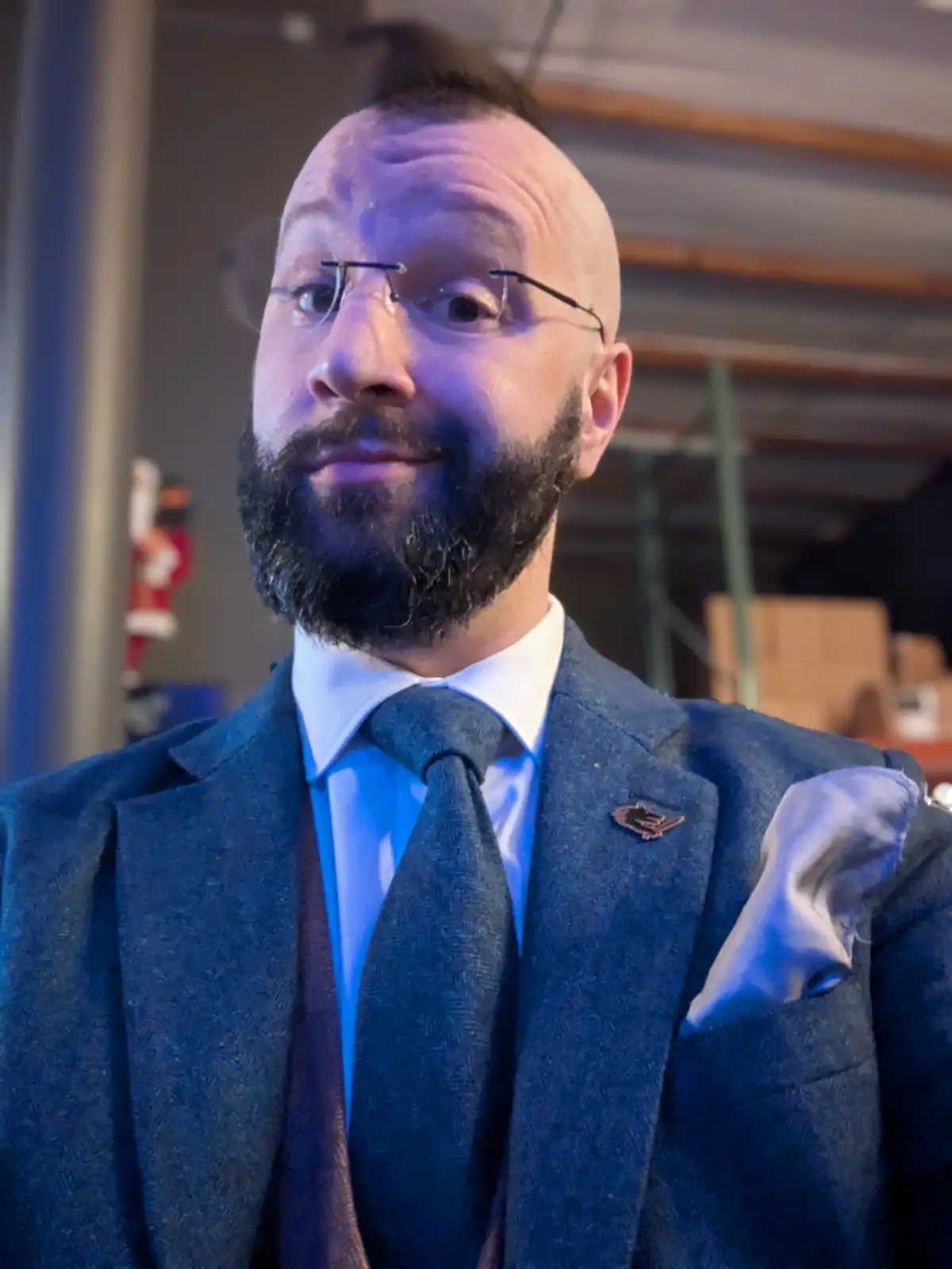 A man with a shaved head, glasses, and a full beard wears a formal blue suit, white shirt, and tie. He has a smug, slightly…