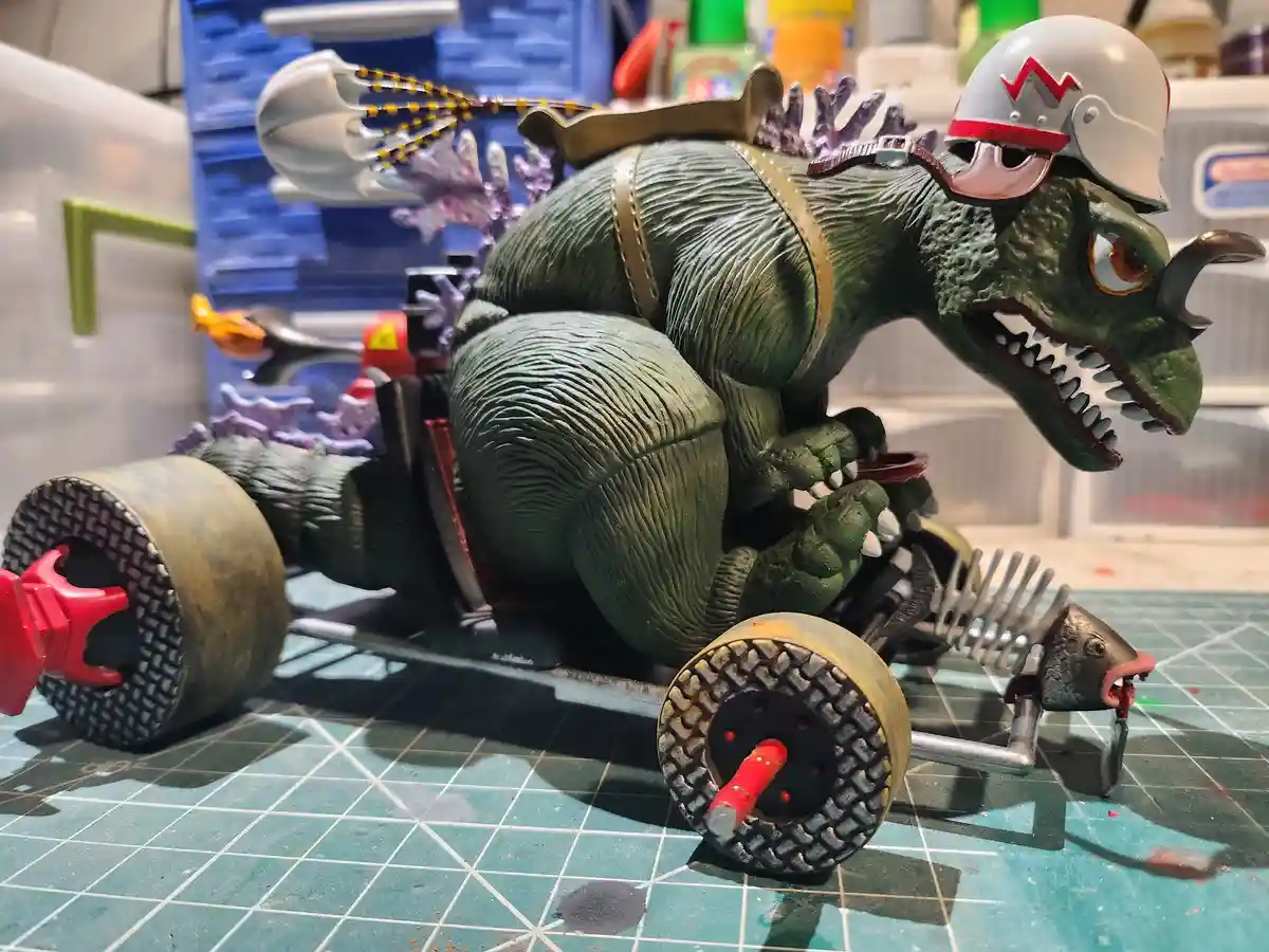 A green Godzilla-like monster figure wearing a white helmet with a red 'M' and goggles, is posed as if driving a…