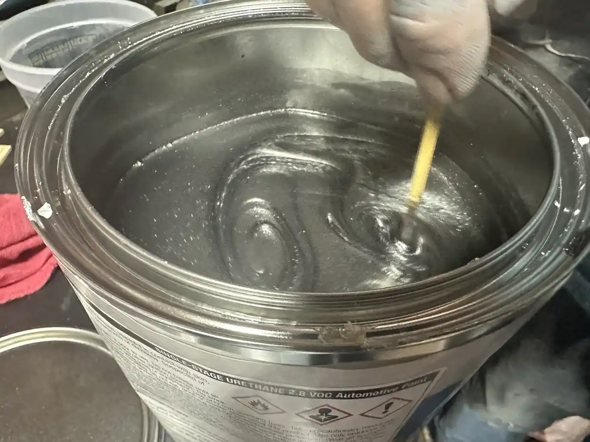 A gloved hand stirs a large metal can of thick, metallic gray automotive paint with a yellow stick. The can's label reads…