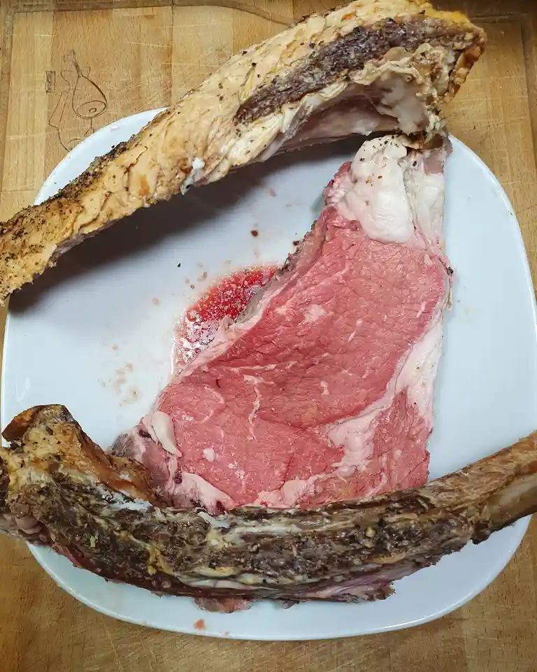 A cooked roast beef with a large bone is sliced open on a white plate, revealing a very pink, rare interior, sitting on a…