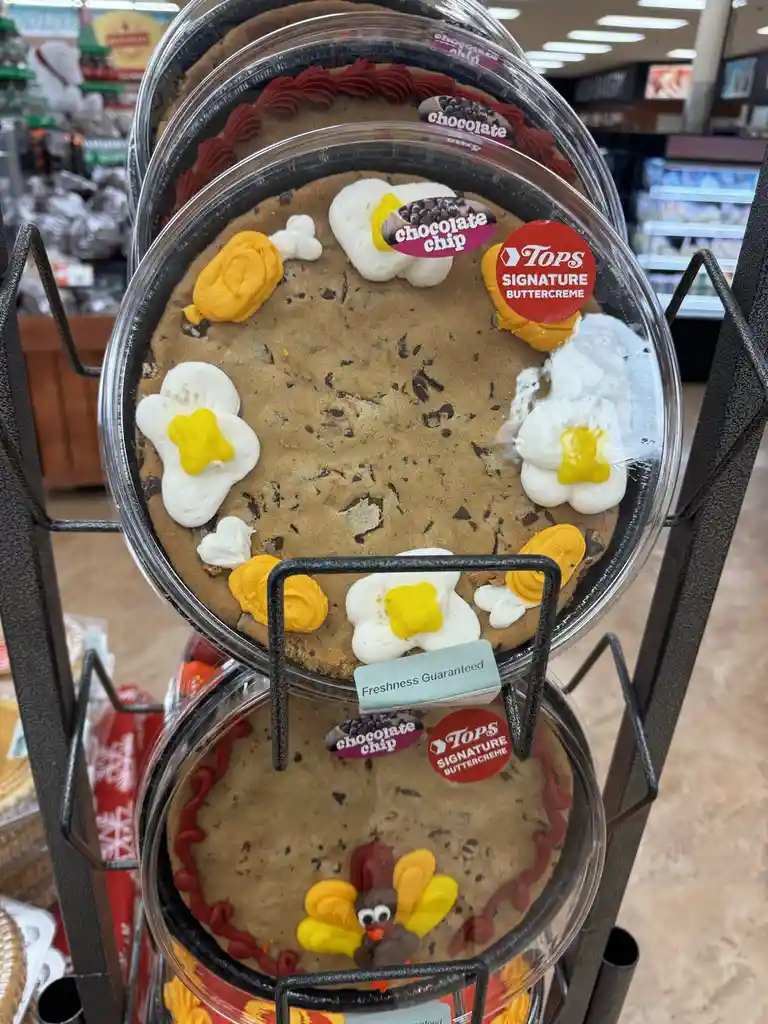A large chocolate chip cookie in a clear plastic container is decorated with white and yellow frosting to resemble fried…