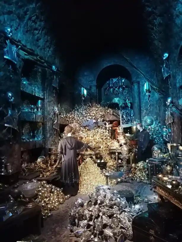 A dimly lit, cavernous room filled with piles of gold and silver trophies, goblets, and ornate objects, with mannequins in…