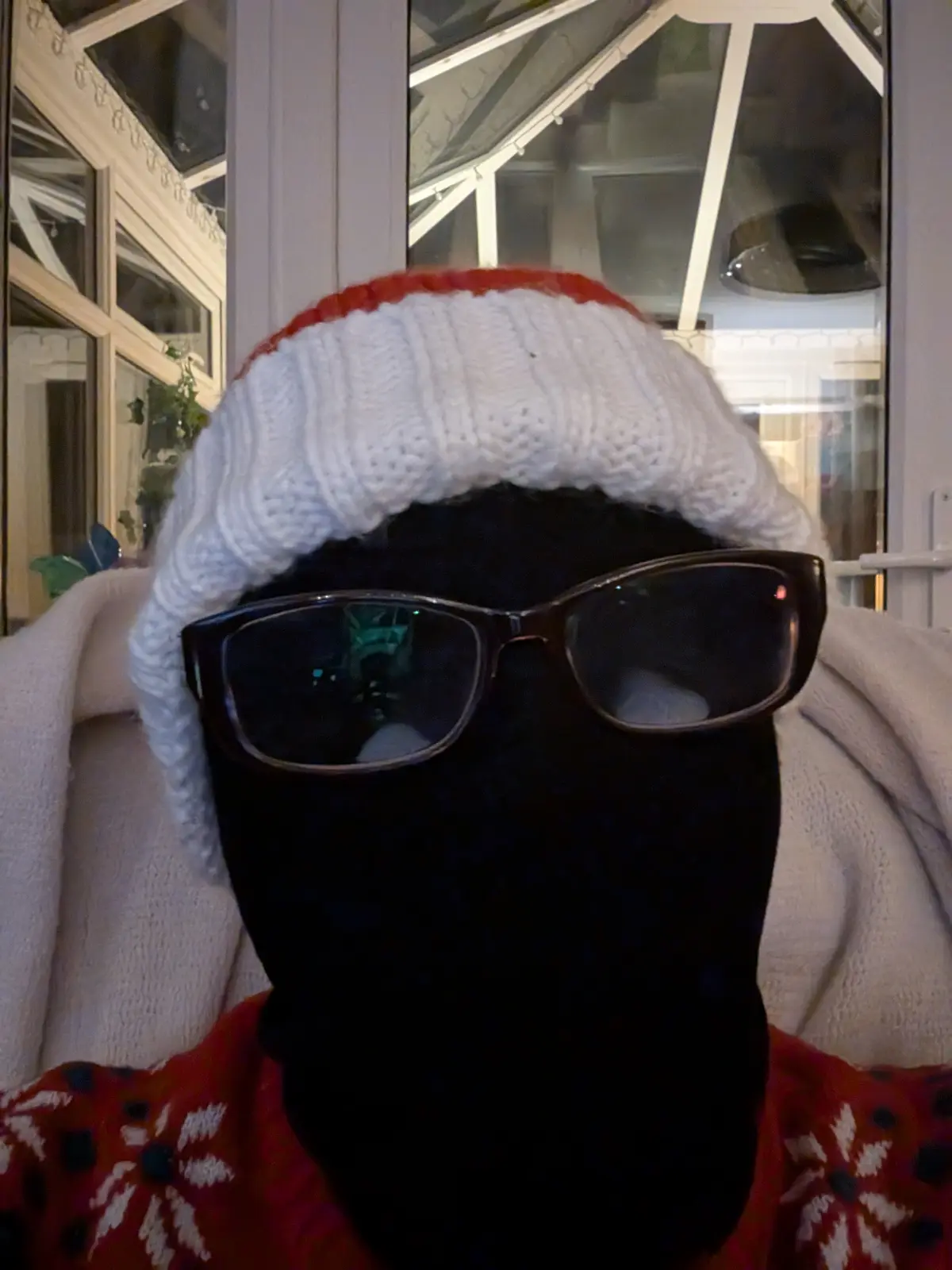 A person is shown from the chest up, wearing a white knit hat with a red brim, a black balaclava covering their entire face…