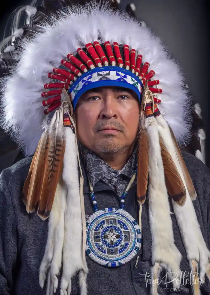 A man with a solemn expression wears a traditional Native American headdress with white feathers, red-tipped adornments, and…