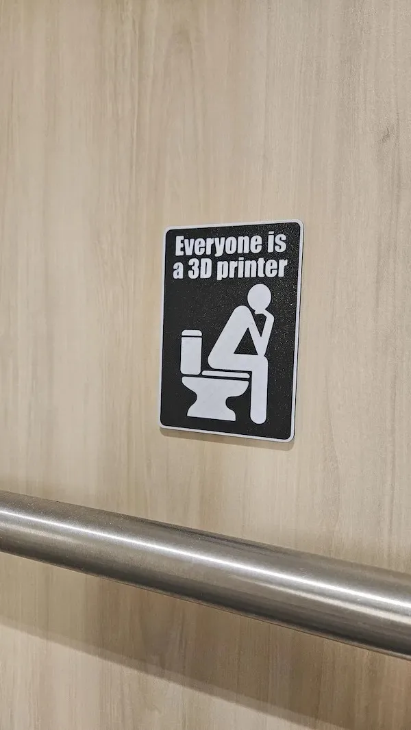 A black-and-white sticker on a light wood-paneled wall depicts a white silhouette of a person sitting on a toilet, with the…