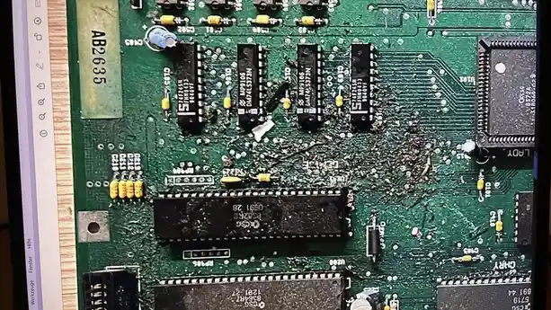 A close-up photo of a severely damaged green circuit board with melted components and burnt residue, overlaid with the text…