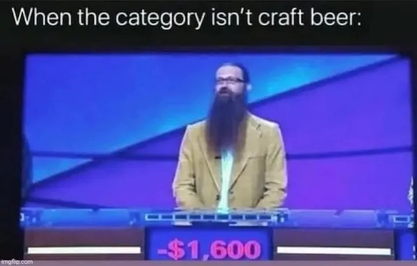 A man with a long beard and glasses, wearing a tan jacket, sits at a game show podium with a blue background. A digital…