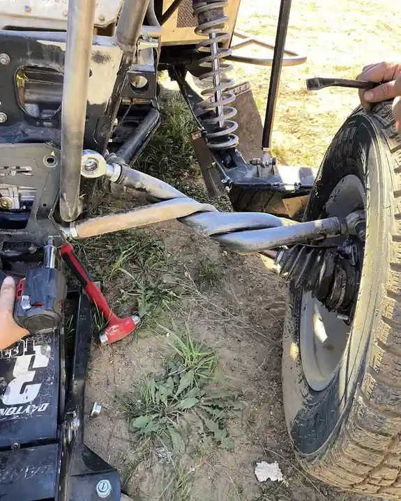 A close-up photo shows a person working on a vehicle's wheel assembly outdoors, with a twisted metal axle shaft visibly…