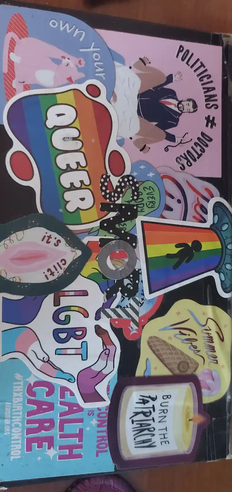 A collage of colorful, overlapping stickers on a surface, featuring phrases like 'own your queeer', 'POLITICIANS ≠ DOCTORS'…