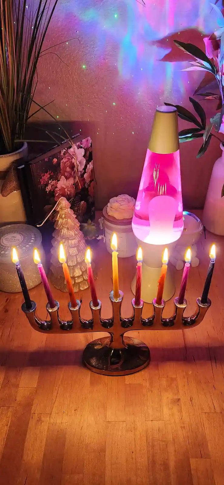 A silver menorah with all nine candles lit sits on a wooden floor, surrounded by decorative items including a pink lava…