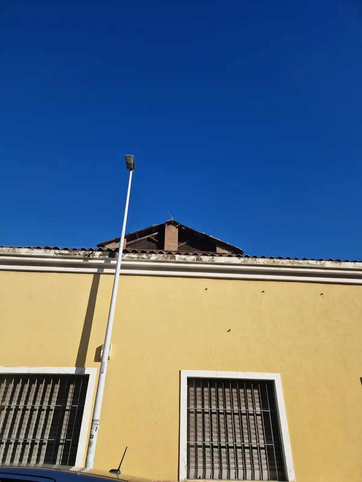 A low-angle photo shows a yellow building with barred windows under a clear blue sky, with a tall white streetlight in the…