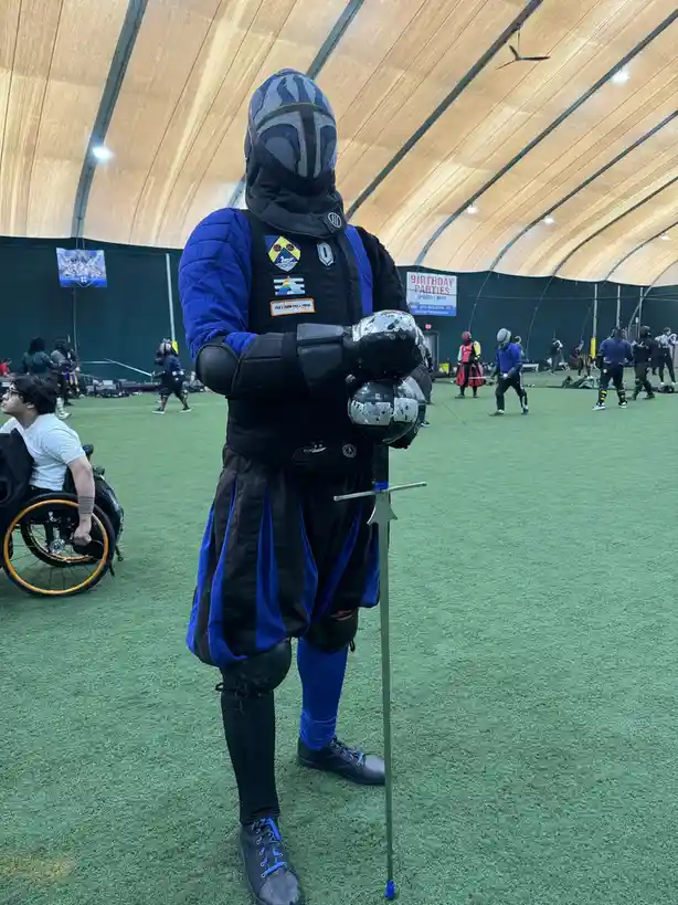A person in full fencing gear, including a mask, blue and black jacket, and protective gear, stands holding a fencing sword…