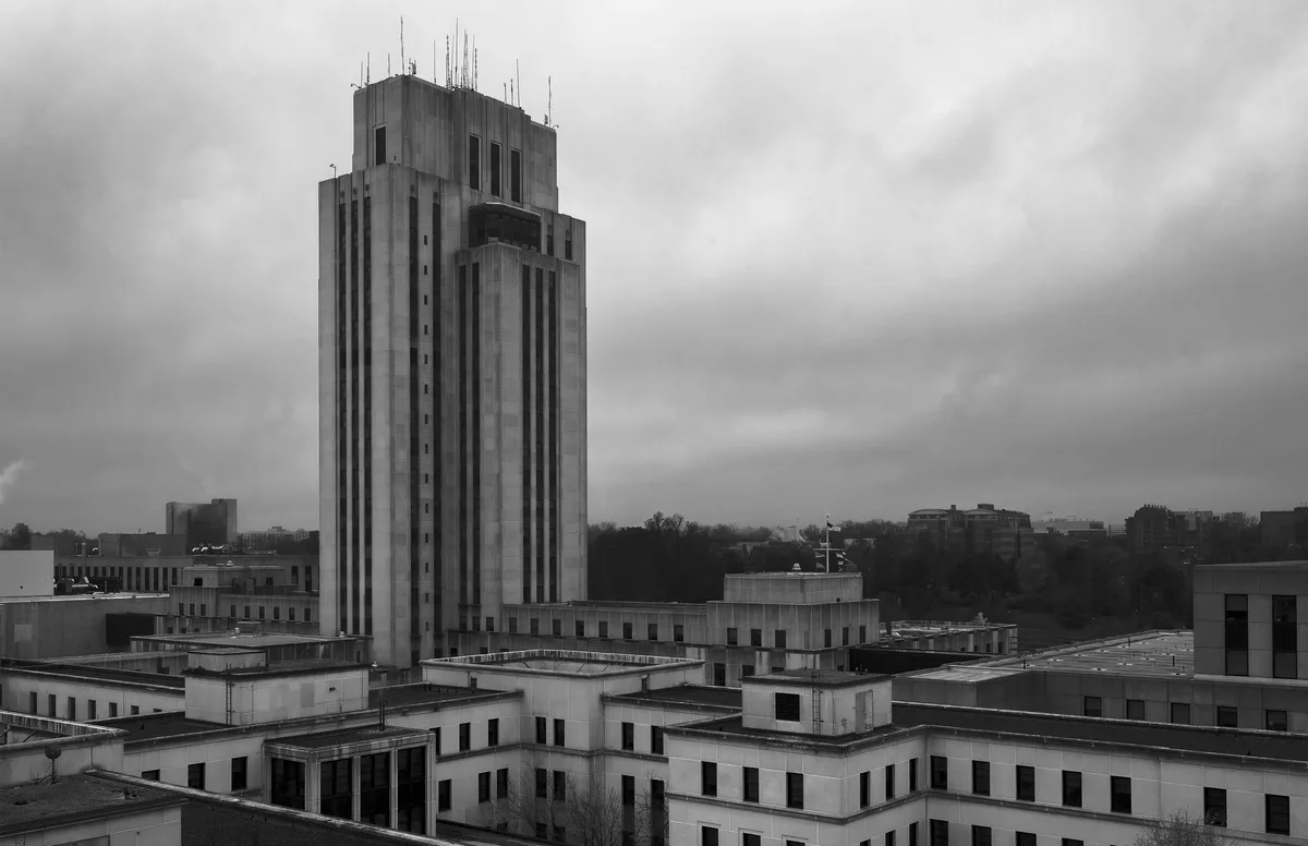 A black-and-white photograph shows a tall, imposing concrete skyscraper dominating a cityscape under a cloudy sky…
